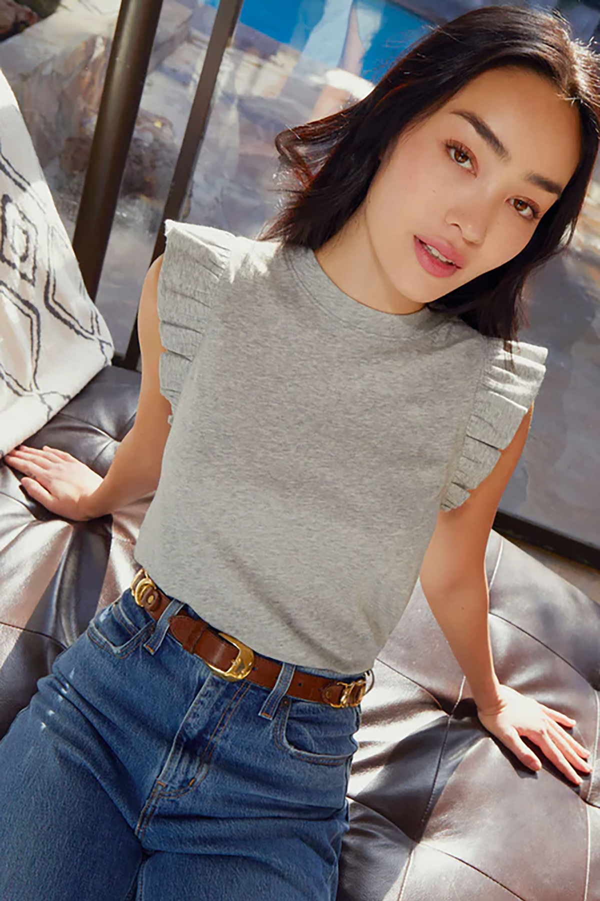 A woman sitting comfortably wearing a gray ruffled flutter sleeve tank top with a belt.