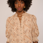 A woman with a large dark curly afro wears a beige and brown patterned blouse with puff sleeves and gold buttons, photographed against a plain light background.