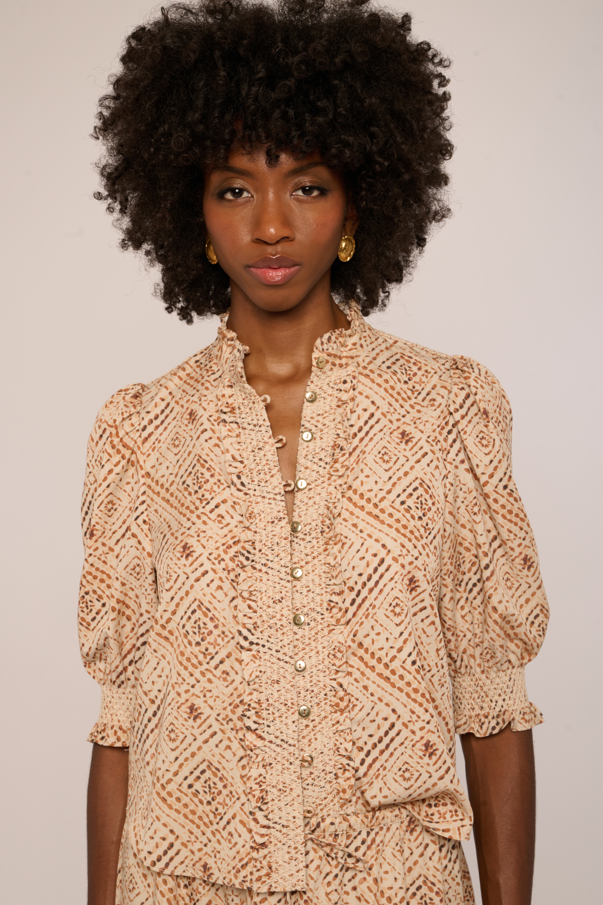 A woman with a large dark curly afro wears a beige and brown patterned blouse with puff sleeves and gold buttons, photographed against a plain light background.