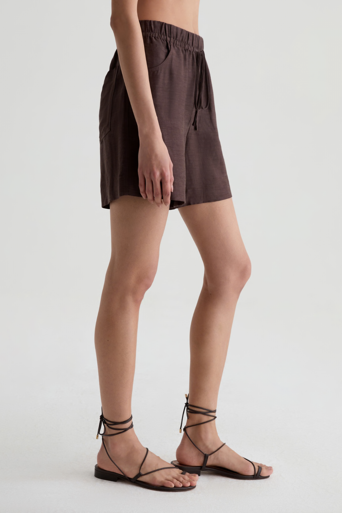 A model wearing dark brown linen shorts with a drawstring waist and black strappy flat sandals, shown from the waist down against a light gray studio background.