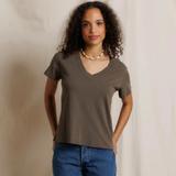 A woman with curly dark hair wearing a taupe brown short-sleeve V-neck t-shirt and blue jeans, standing against a draped beige fabric backdrop.