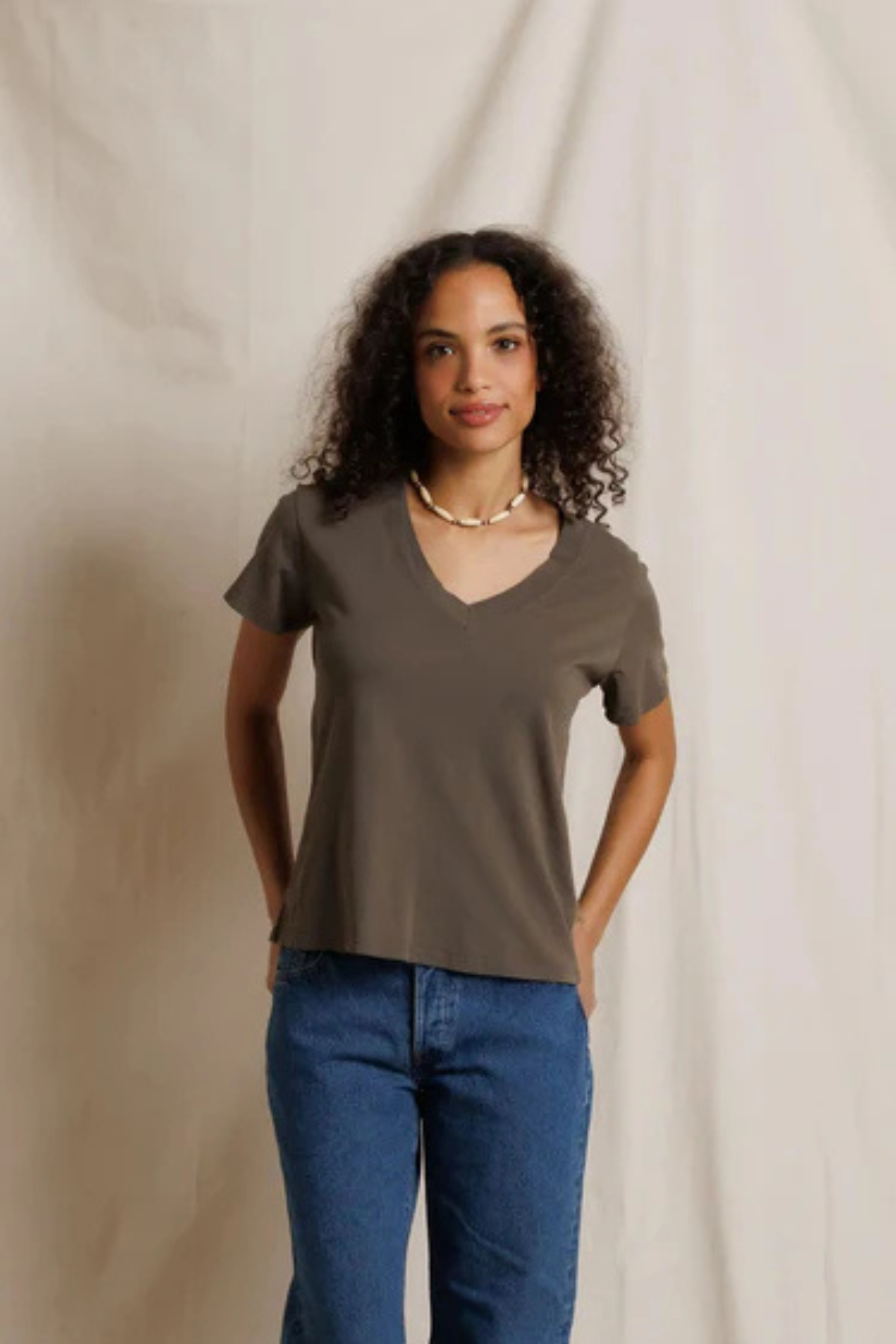 A woman with curly dark hair wearing a taupe brown short-sleeve V-neck t-shirt and blue jeans, standing against a draped beige fabric backdrop.