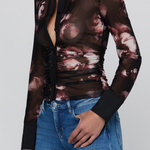 A woman wearing a sheer long-sleeve button-up blouse with a black collar and cuffs, featuring a dark brown and dusty rose floral print, paired with blue jeans against a light gray background.