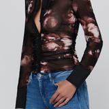 A woman wearing a sheer long-sleeve button-up blouse with a black collar and cuffs, featuring a dark brown and dusty rose floral print, paired with blue jeans against a light gray background.