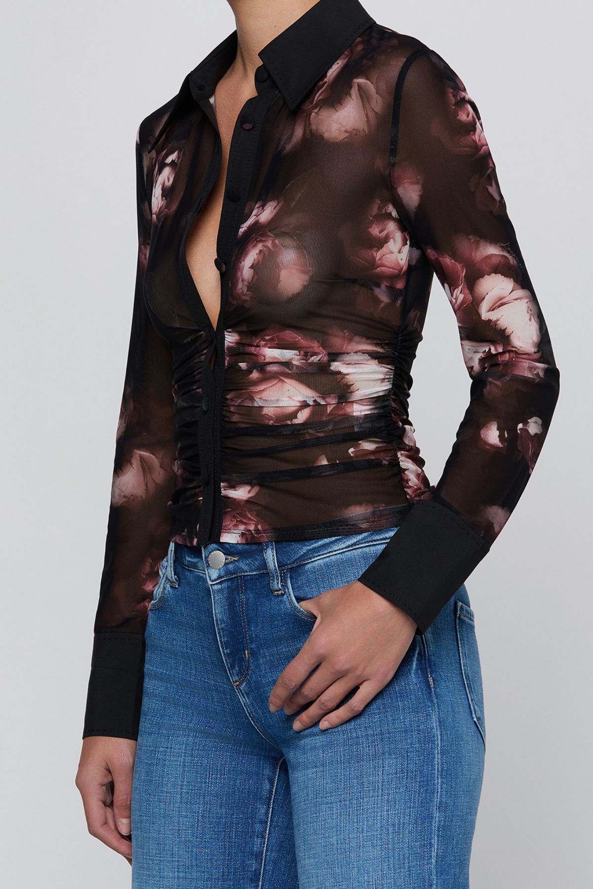 A woman wearing a sheer long-sleeve button-up blouse with a black collar and cuffs, featuring a dark brown and dusty rose floral print, paired with blue jeans against a light gray background.