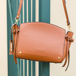 A tan leather shoulder bag with gold-tone hardware and tassel details, hanging in front of a teal-green slatted door.