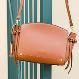 A tan leather shoulder bag with gold-tone hardware and tassel details, hanging in front of a teal-green slatted door.
