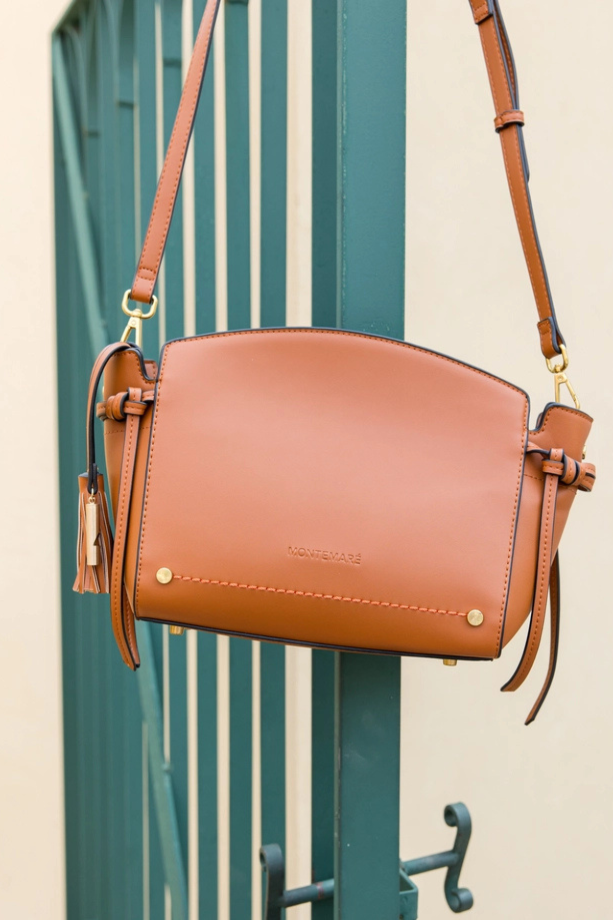 A tan leather shoulder bag with gold-tone hardware and tassel details, hanging in front of a teal-green slatted door.