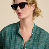 A woman with blonde hair pulled back wears black cat-eye sunglasses and a green plaid button-up shirt against a solid beige background.