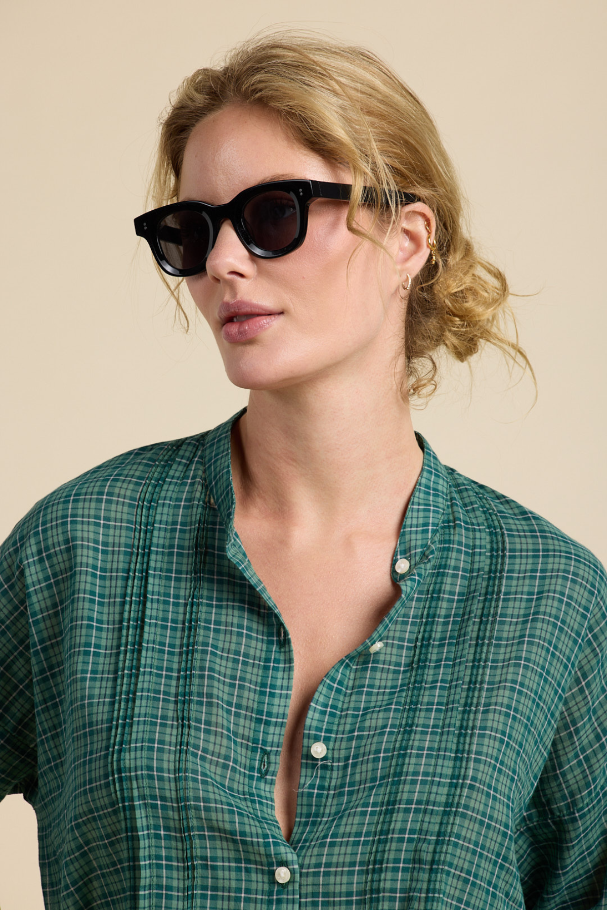 A woman with blonde hair pulled back wears black cat-eye sunglasses and a green plaid button-up shirt against a solid beige background.