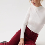 A woman sitting on a white studio floor wearing a fitted white mock-neck long-sleeve top and high-waisted burgundy corduroy trousers.