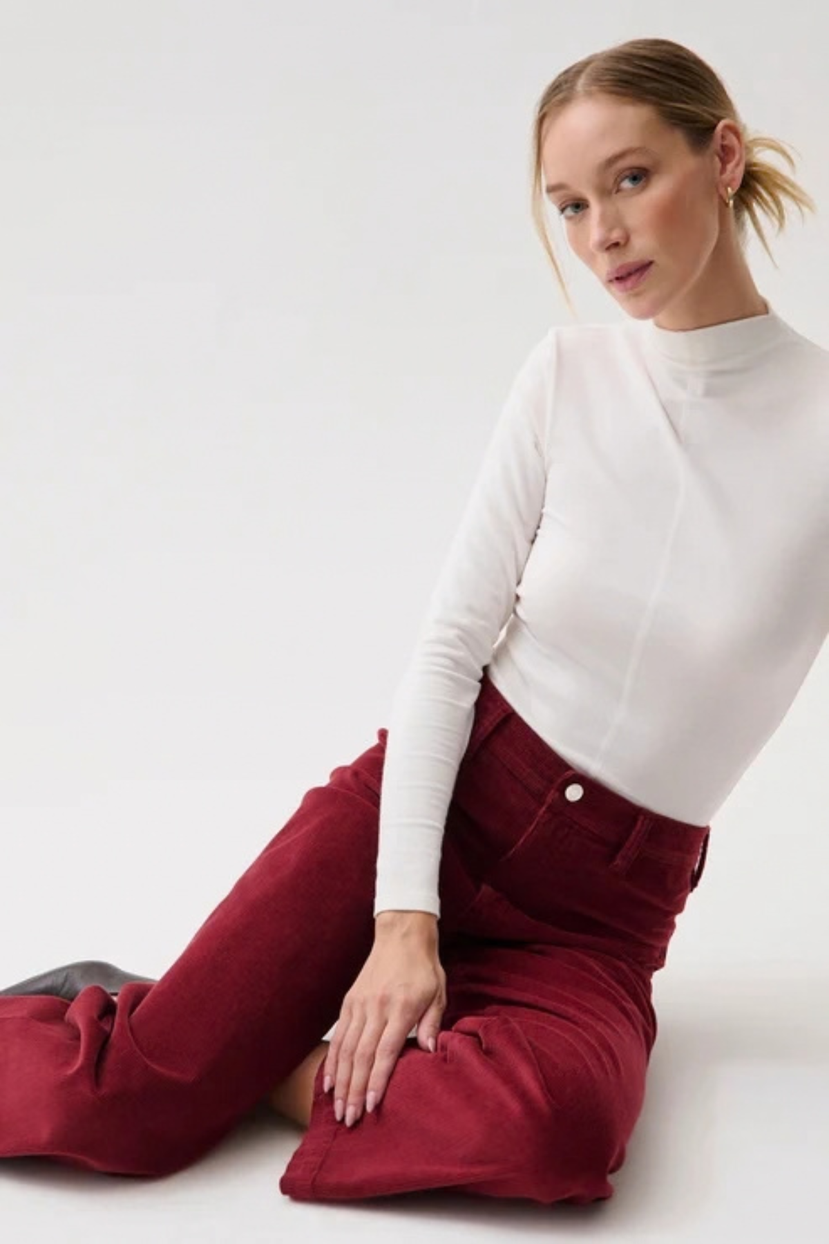 A woman sitting on a white studio floor wearing a fitted white mock-neck long-sleeve top and high-waisted burgundy corduroy trousers.