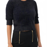 A woman wearing a dark navy fuzzy cropped sweater with three-quarter sleeves and black pants with gold zipper details, photographed against a plain white background.