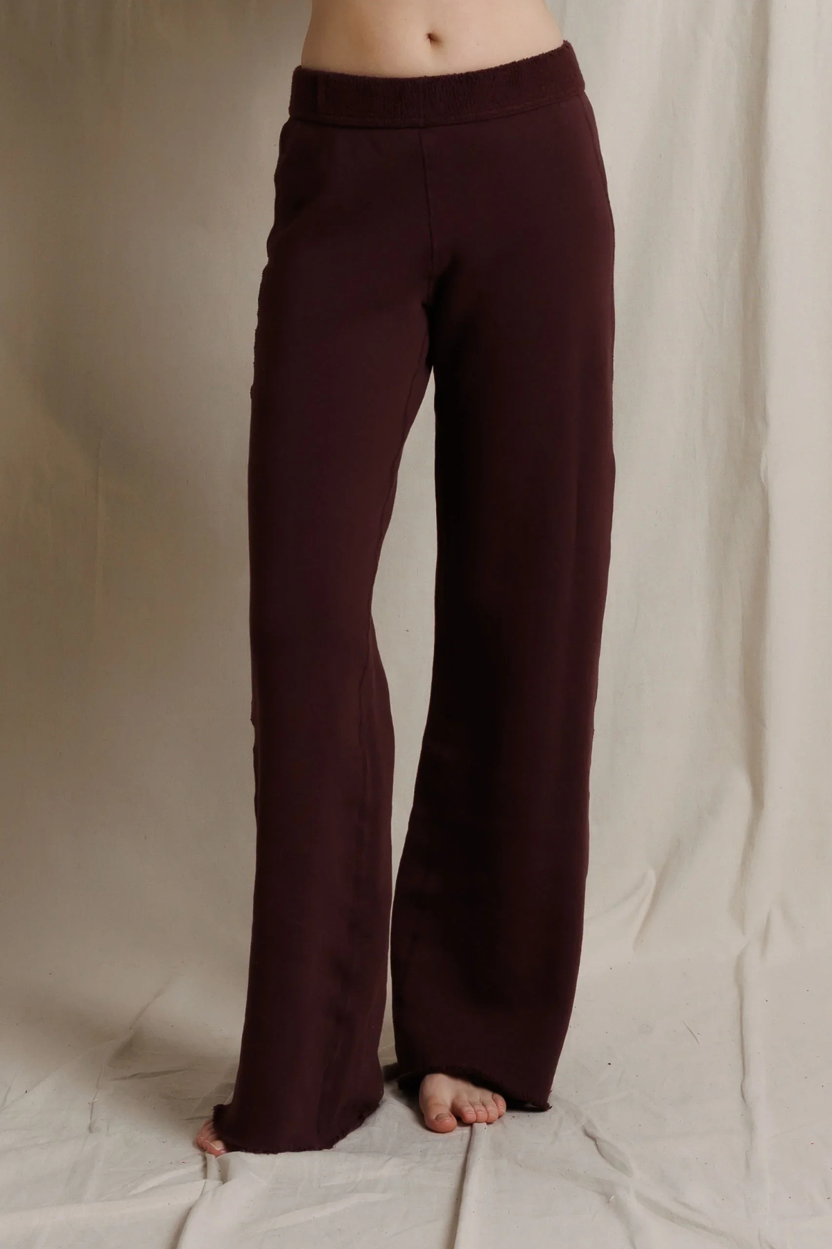 A person wearing dark burgundy wide-leg pants, shown from the waist down against a draped off-white fabric backdrop.