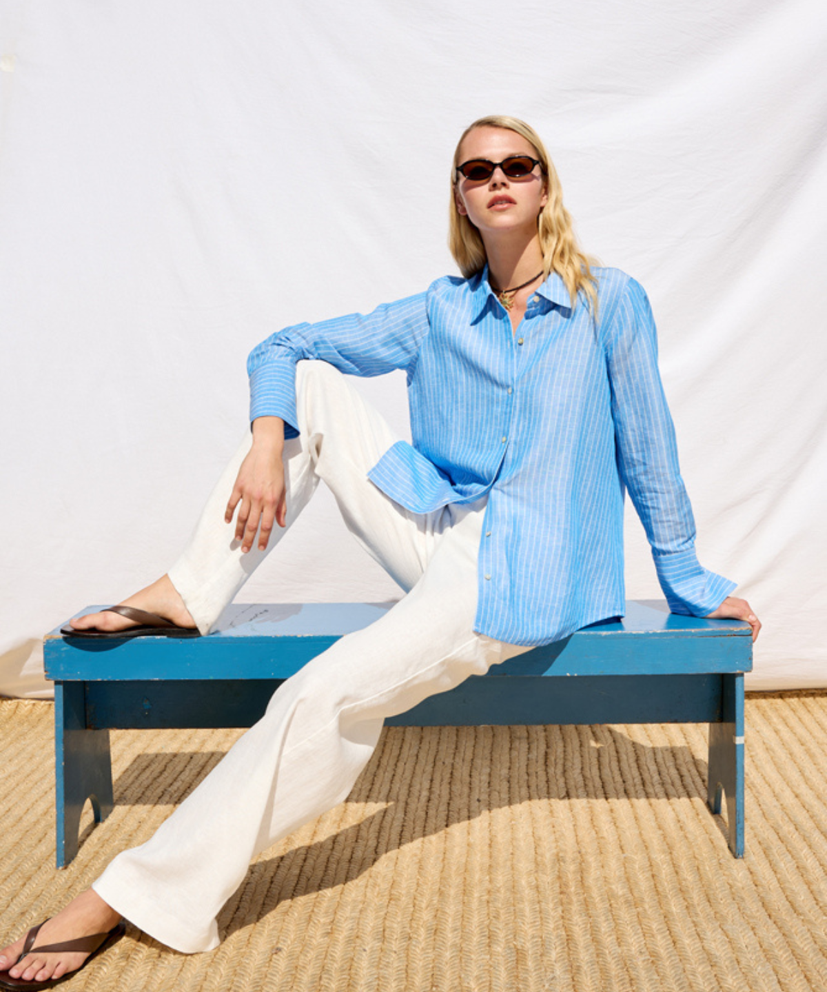 Woman in a blue shirt and white pants sitting on a blue bench against a white background
