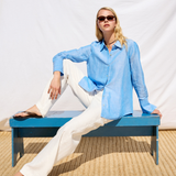 Woman in a blue shirt and white pants sitting on a blue bench against a white background