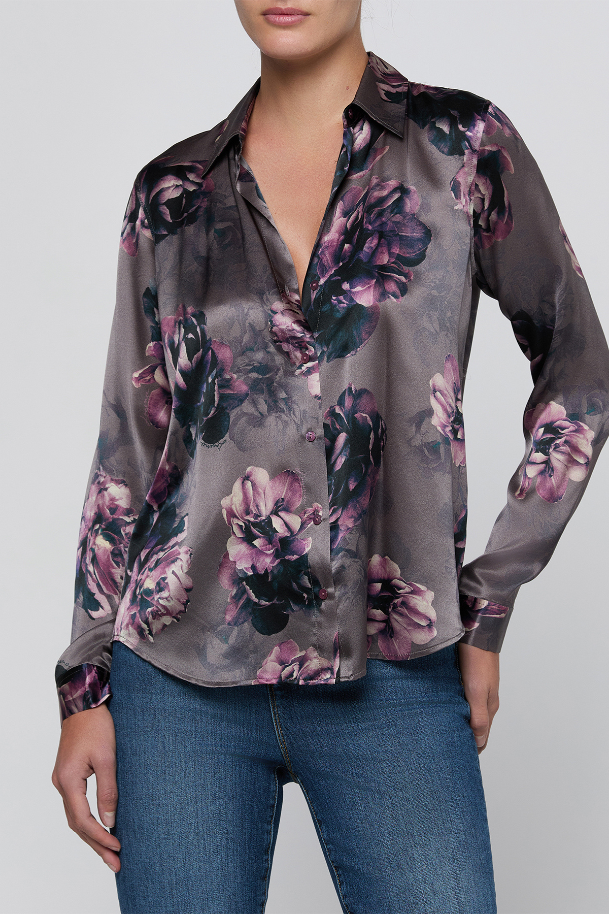 A woman wearing a long-sleeve satin button-up blouse with a dark gray base and a large floral print in purple, pink, and black, paired with blue jeans against a light gray studio background.