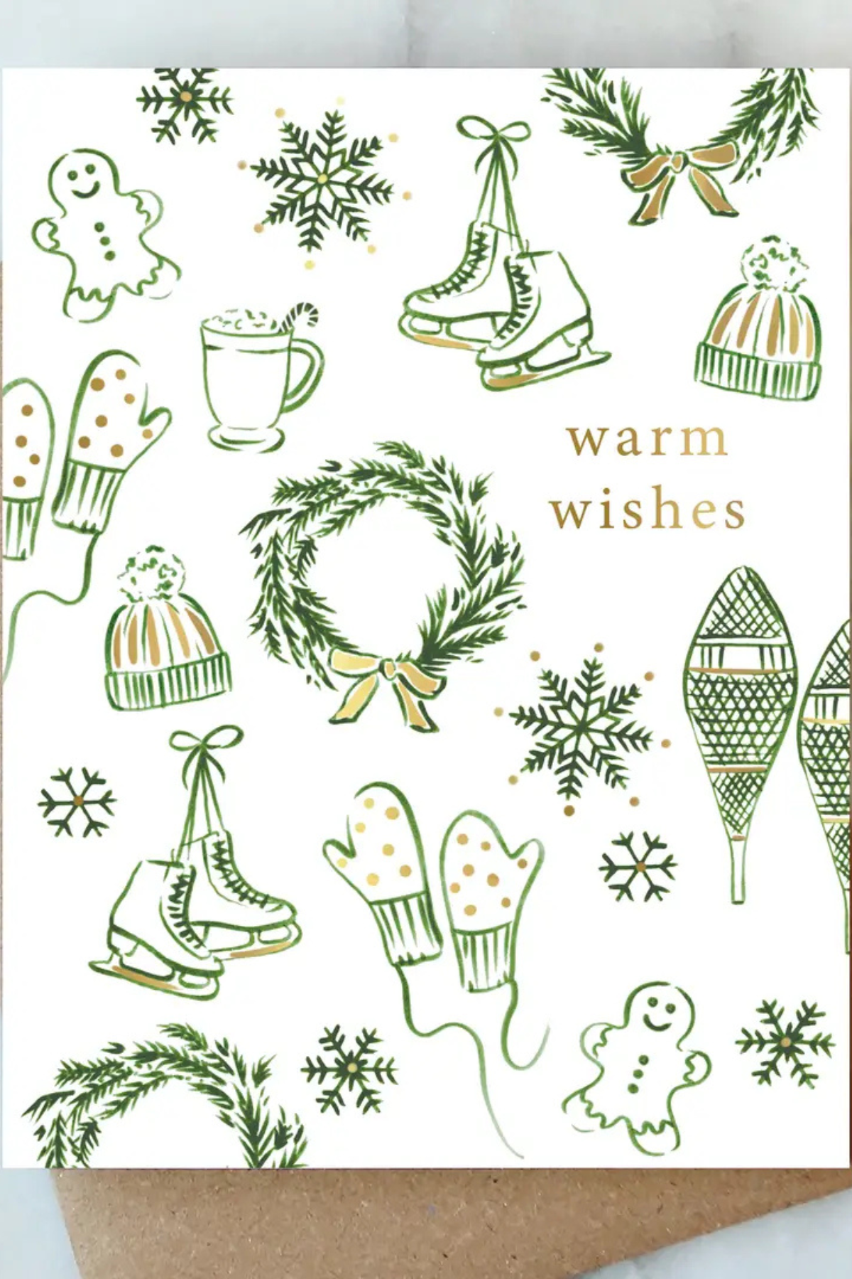 A white holiday greeting card with green line drawings of snowflakes, wreaths, ice skates, mittens, snow boots, gingerbread cookies, and pine branches, with the words 'warm wishes' in gold. A brown kraft envelope is partially visible underneath.