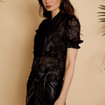 A woman modeling a black sheer lace short-sleeve blouse with a dark floral pattern, paired with a black faux-leather mini skirt, set against a warm beige background with decorative palm leaves.