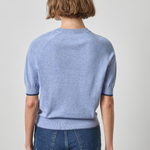 A model shown from the back wearing a light blue short-sleeve knit sweater and dark blue jeans against a plain light gray studio background.