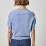 A model shown from the back wearing a light blue short-sleeve knit sweater and dark blue jeans against a plain light gray studio background.