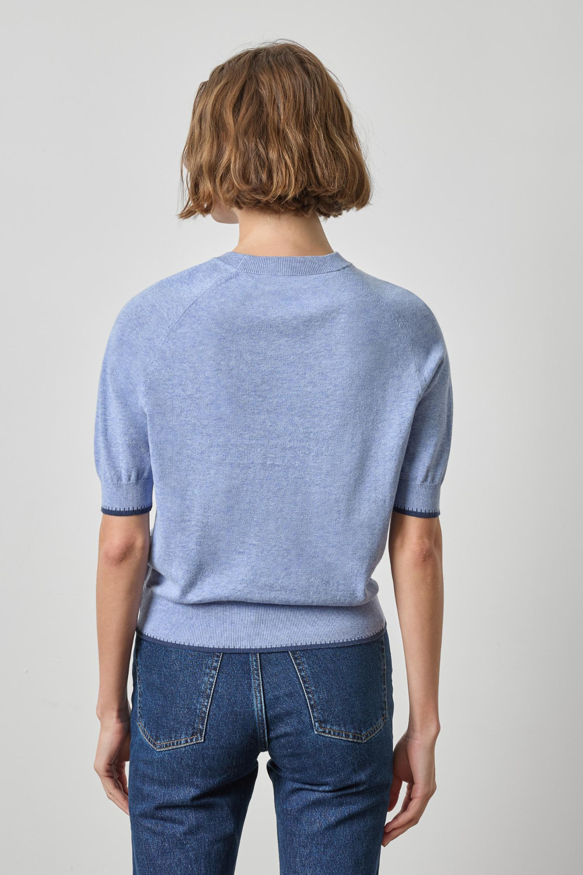 A model shown from the back wearing a light blue short-sleeve knit sweater and dark blue jeans against a plain light gray studio background.