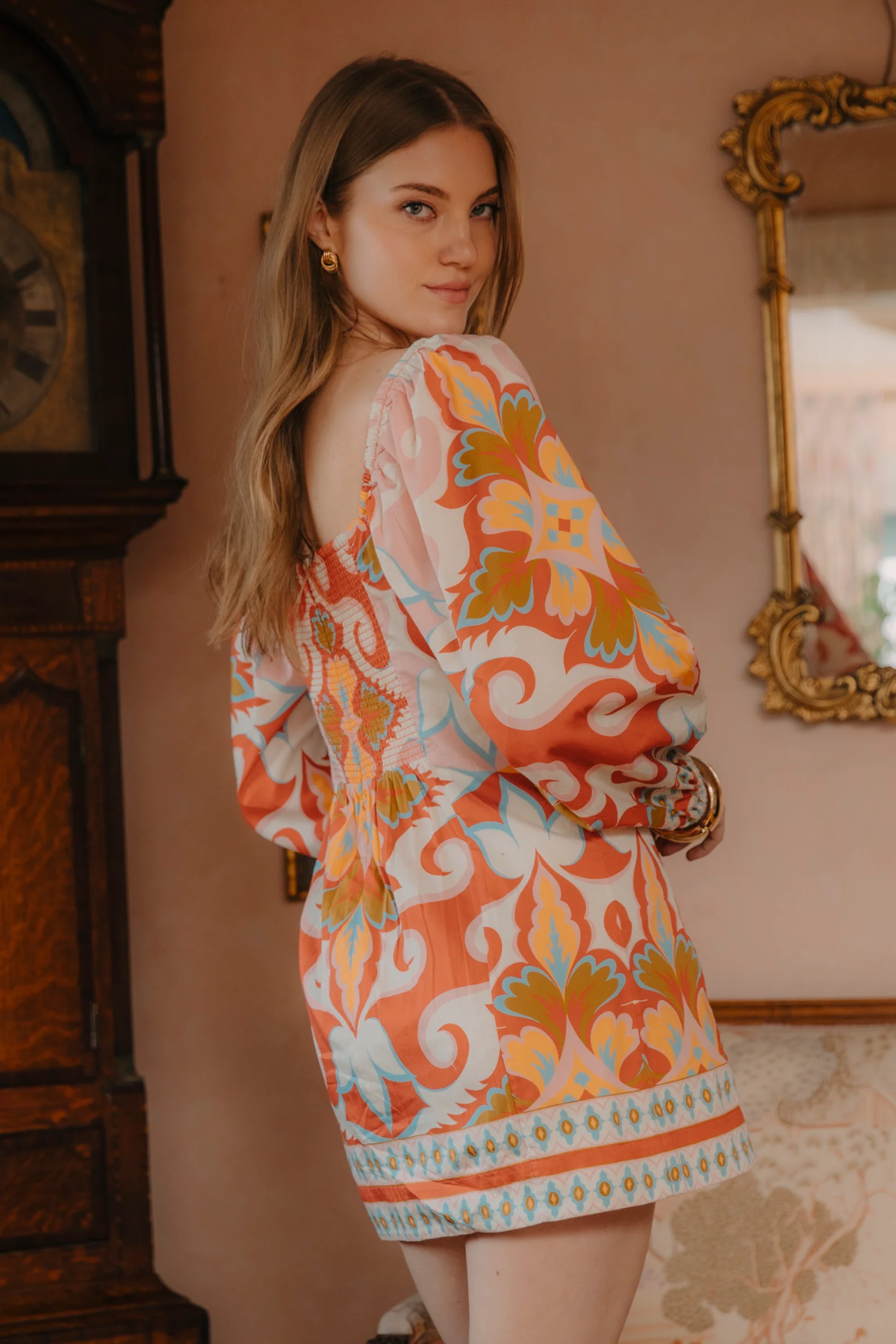 Woman wearing a colorful patterned dress in an indoor setting with a mirror and clock.
