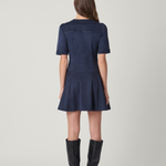 A woman viewed from the back wearing a short-sleeve navy blue mini dress and black knee-high boots, standing against a light gray studio background.