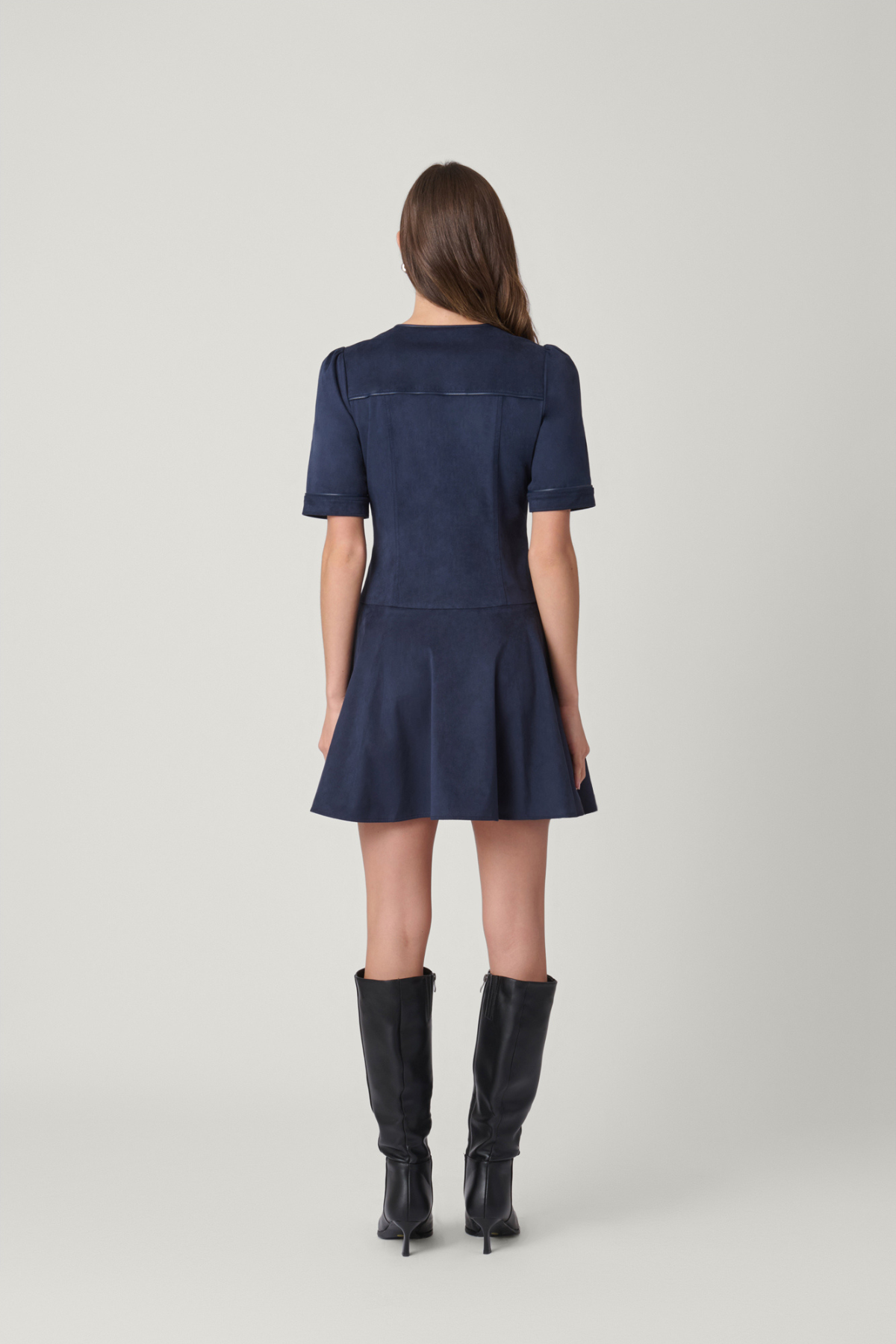 A woman viewed from the back wearing a short-sleeve navy blue mini dress and black knee-high boots, standing against a light gray studio background.