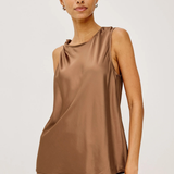 A woman modeling a sleeveless taupe-brown satin top with a draped neckline, paired with a matching satin skirt, against a plain light gray studio background.