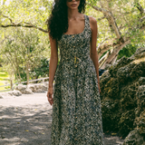 A woman wearing a sleeveless black and beige animal print midi dress with a tie waist, standing outdoors on a gravel path surrounded by trees and rocks.