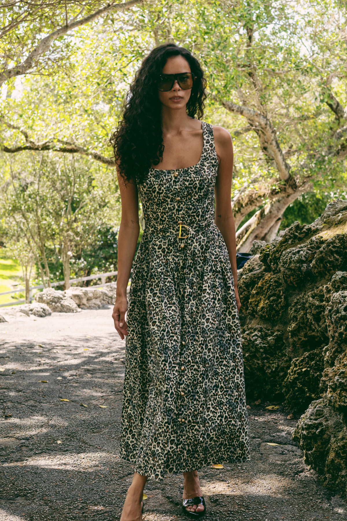 A woman wearing a sleeveless black and beige animal print midi dress with a tie waist, standing outdoors on a gravel path surrounded by trees and rocks.