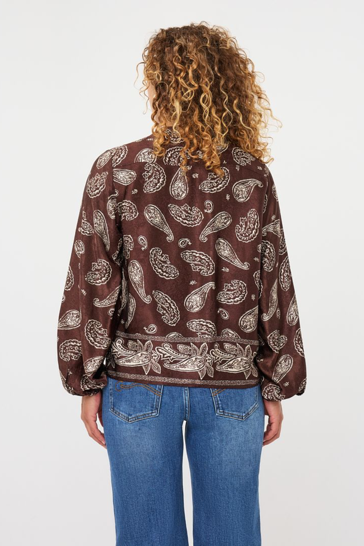 A person viewed from the back wearing a brown long-sleeve blouse with a beige paisley pattern, paired with blue jeans, against a plain white background.