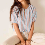 Woman wearing a striped blouse sitting on a woven stool against a plain background