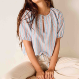 Woman wearing a striped blouse sitting on a woven stool against a plain background